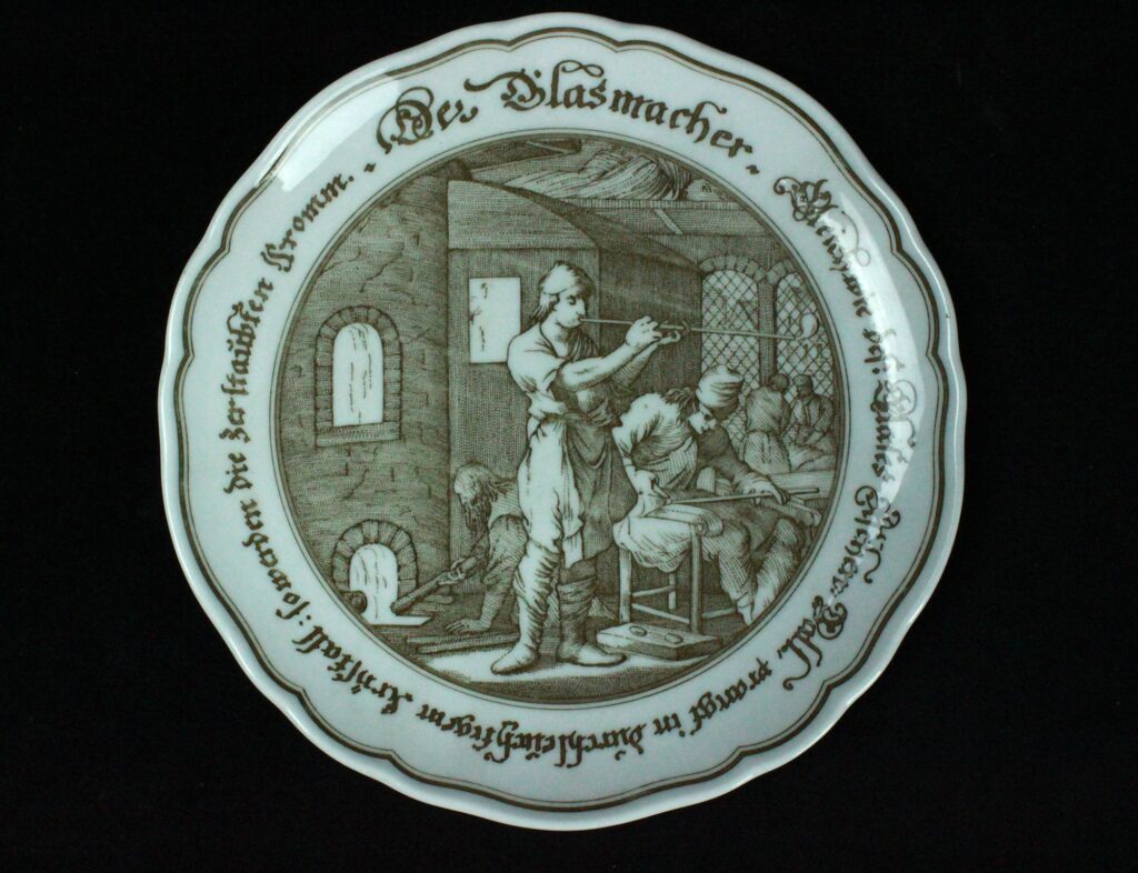 Der Coupe-Kuchenteller hat ein Dekor in Schiebbildtechnik mit der Abbildung aus Jost Ammanns mittelalterlichem Werk über Handwerke. Konkret findet sich "Der Glasmacher" abgebildet mit dem Spruchband "Gleich wie des Glases Aschenball prangt in durchleuchtigem Krystall. So werden die zerstaubten fromm"